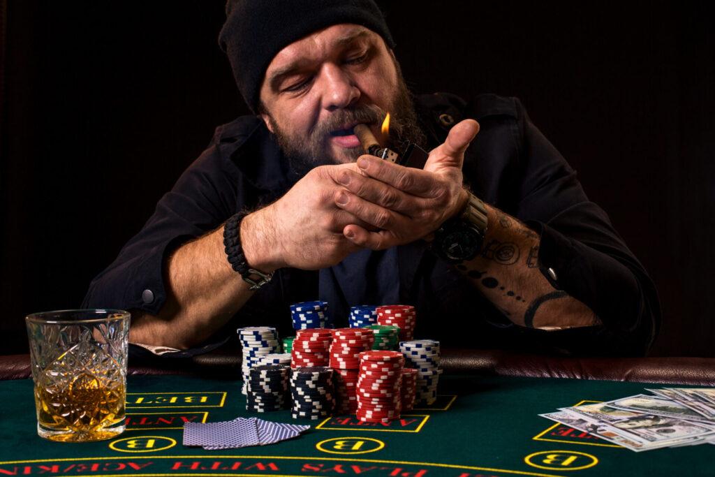 Vousatý muž s doutníkem a sklenicí sedí u pokerového stolu v kasinu. Hraje karty a ruletu, na zeleném stole jsou žetony, karty a peníze. Celý prostor je zahalen kouřem z doutníků.