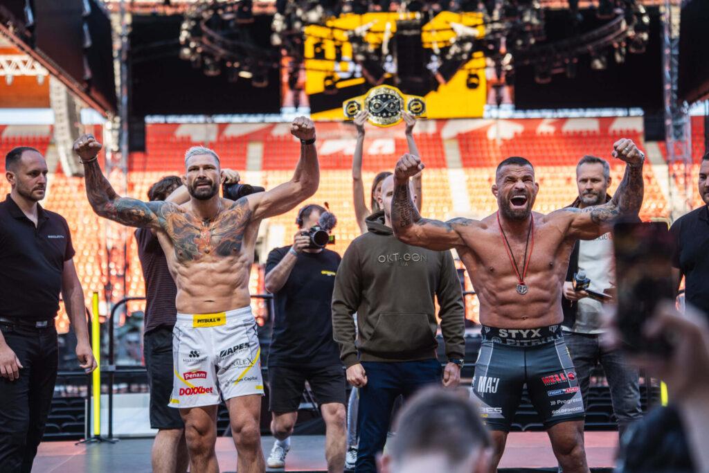 Attila Végh a Karlos Vémola pózují se zdviženými pažemi na pódiu při vážení před zápasem Oktagon MMA v Edenu. V pozadí je pás šampiona a prázdné tribuny stadionu. Atmosféra je napjatá a plná očekávání.
